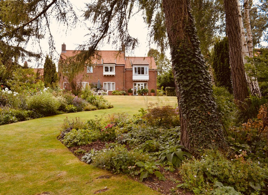 A well-maintained garden scene featuring a lush, green lawn with neatly trimmed grass and varied flower beds filled with blooming plants and shrubs. The garden is shaded by large, mature trees with te